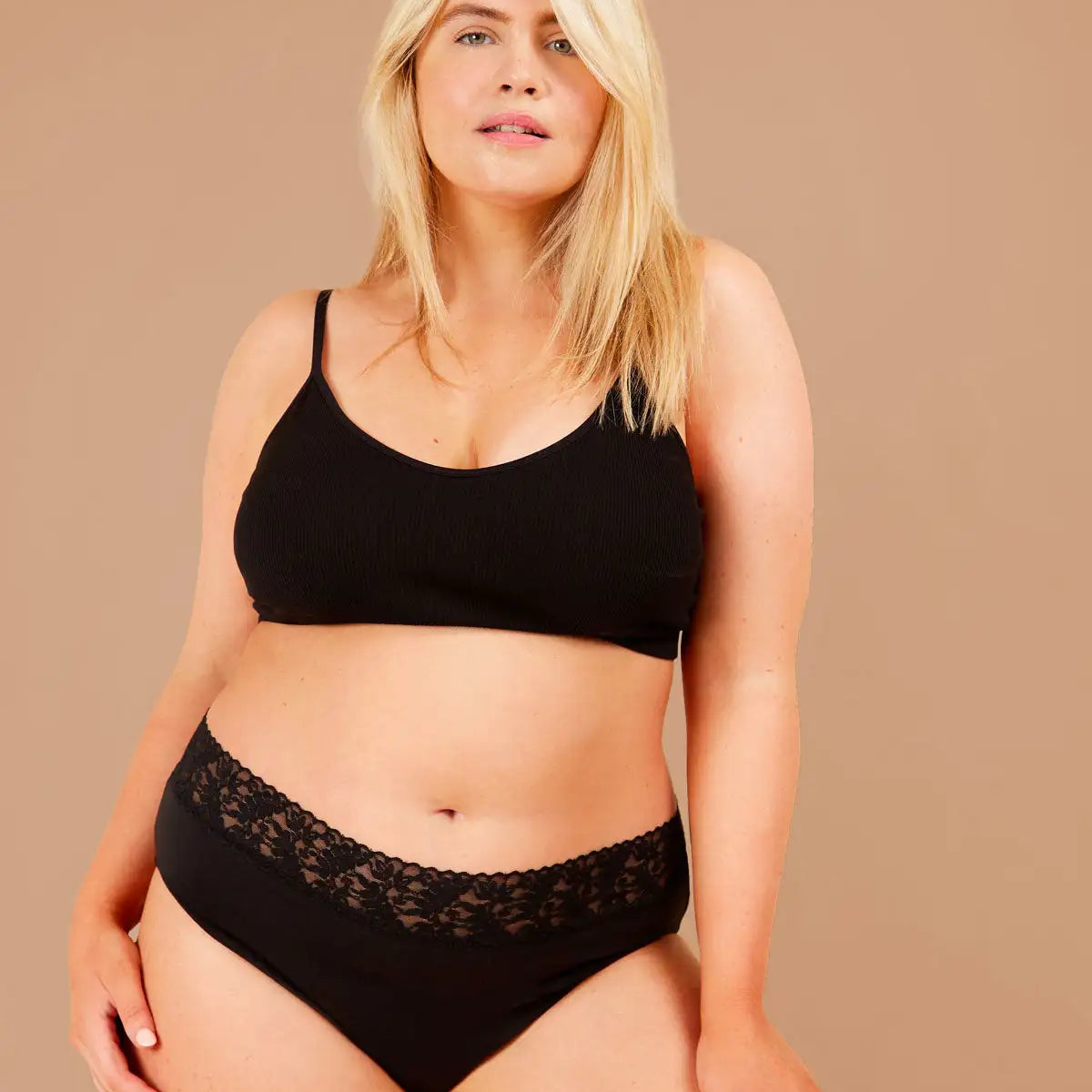 Blonde woman standing confidently, wearing black period pants with lacey floral trim gazes directly at the camera.