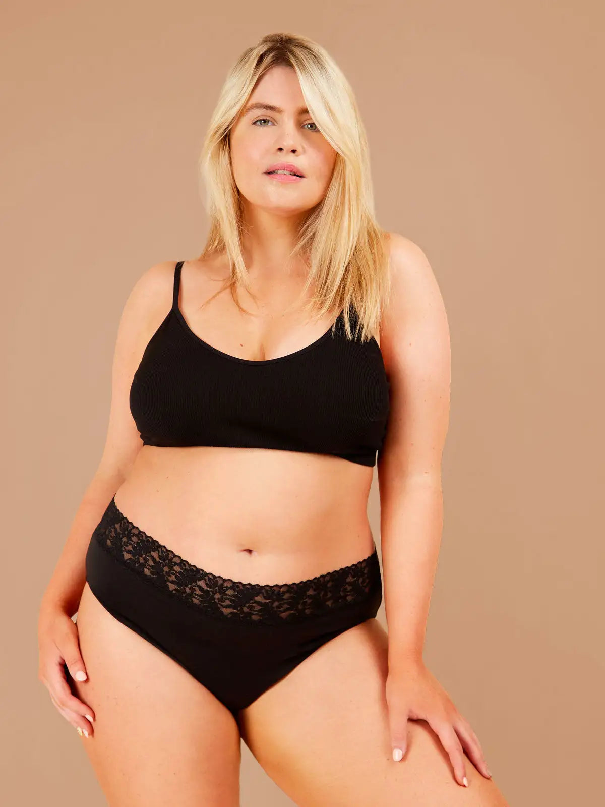 Blonde woman standing confidently, wearing black period pants with lacey floral trim gazes directly at the camera.